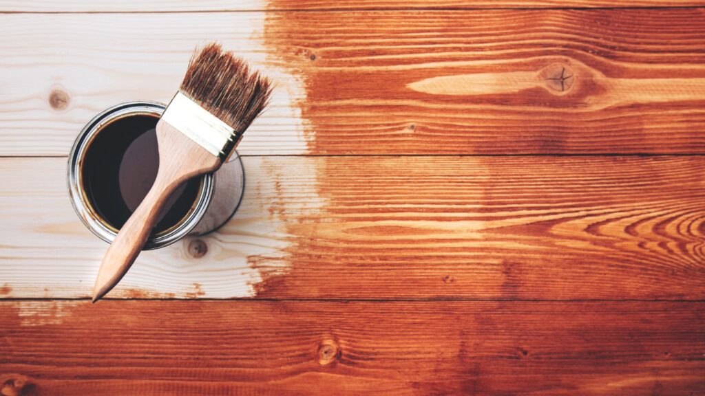A paintbrush resting on an open can of wood stain, partially applied to a wooden surface